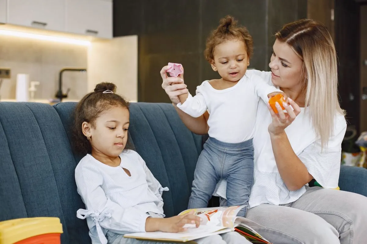 Una babysitter gioca con le costruzioni e sfoglia un libro insieme a due bambini sul divano di casa