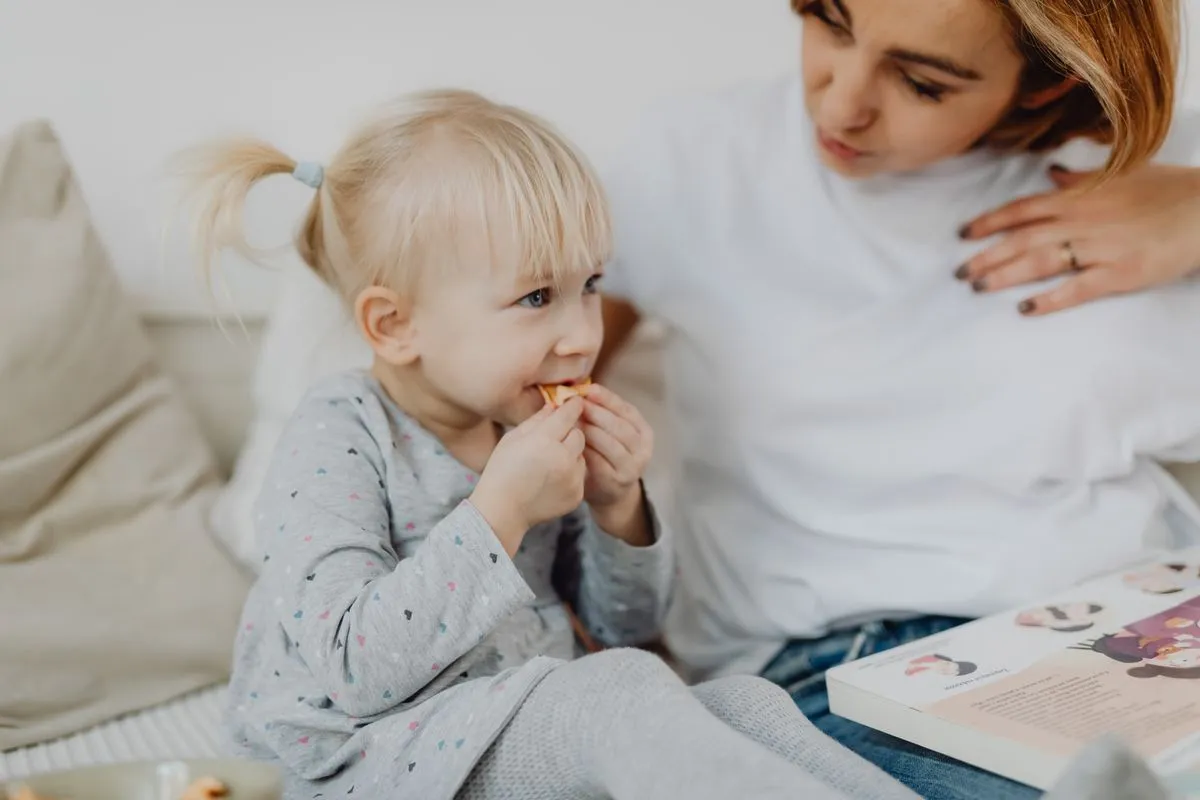 Una tata accompagna la merenda di una bambina seduta sul divano, con un libro illustrato accanto