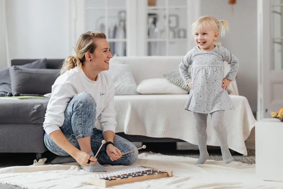 Una babysitter suona uno xilofono sul tappeto mentre una bambina sorride e balla in salotto